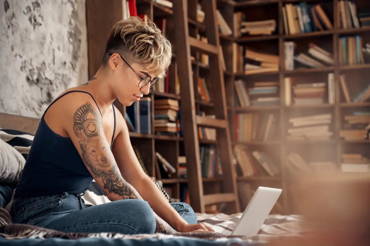 Person with a tattoo working on a laptop in a cozy room filled with books, representing ideas for a side hustle.