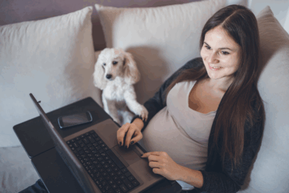 Pregnant woman smiling while working on a laptop from the couch with her dog beside her, representing a flexible Amazon side hustle.
