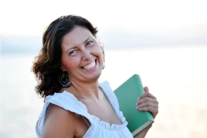 Smiling woman in her 40s holding a notebook at the beach, symbolizing confidence and freedom through side hustles for women over 40.