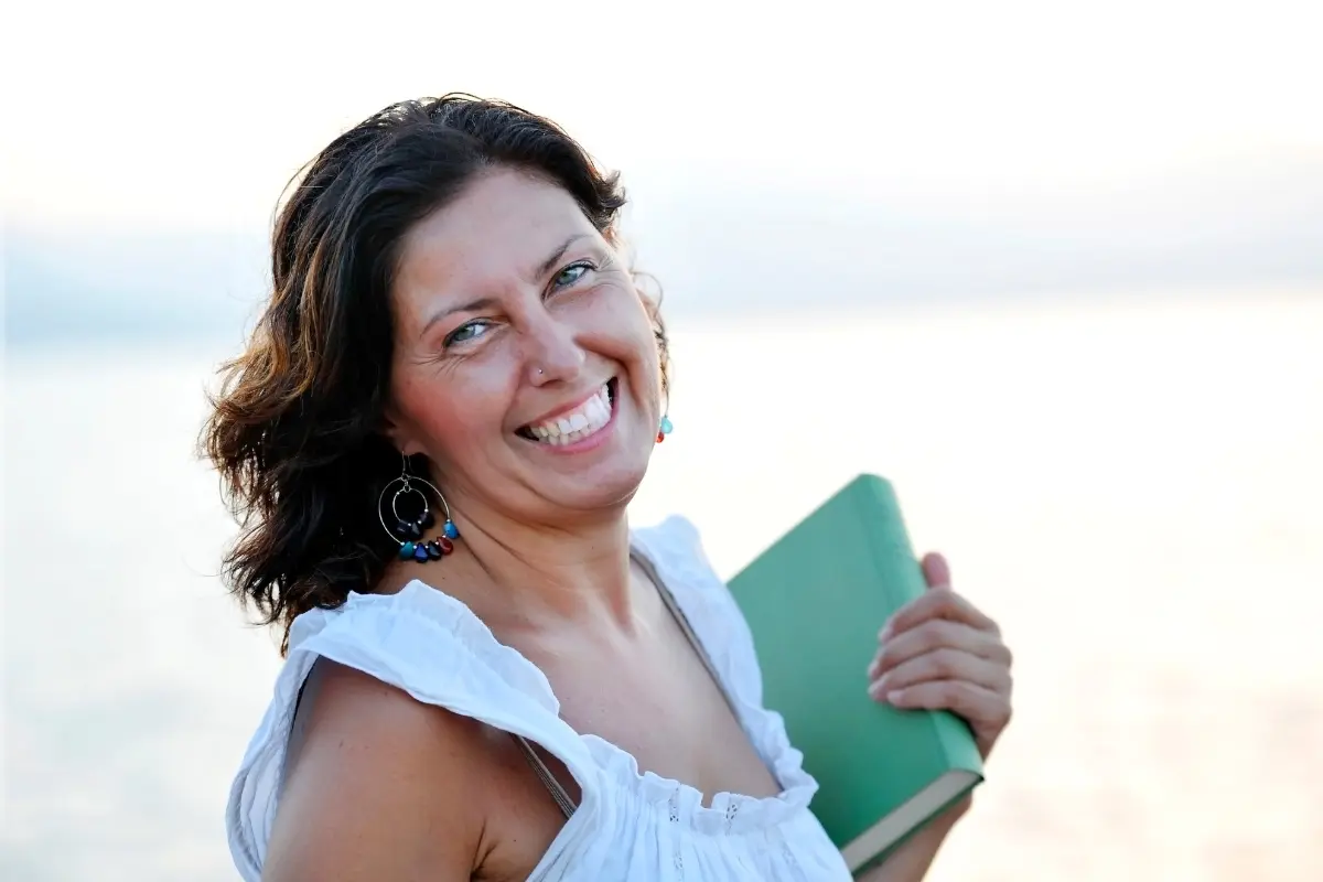 Smiling woman in her 40s holding a notebook at the beach, symbolizing confidence and freedom through side hustles for women over 40.