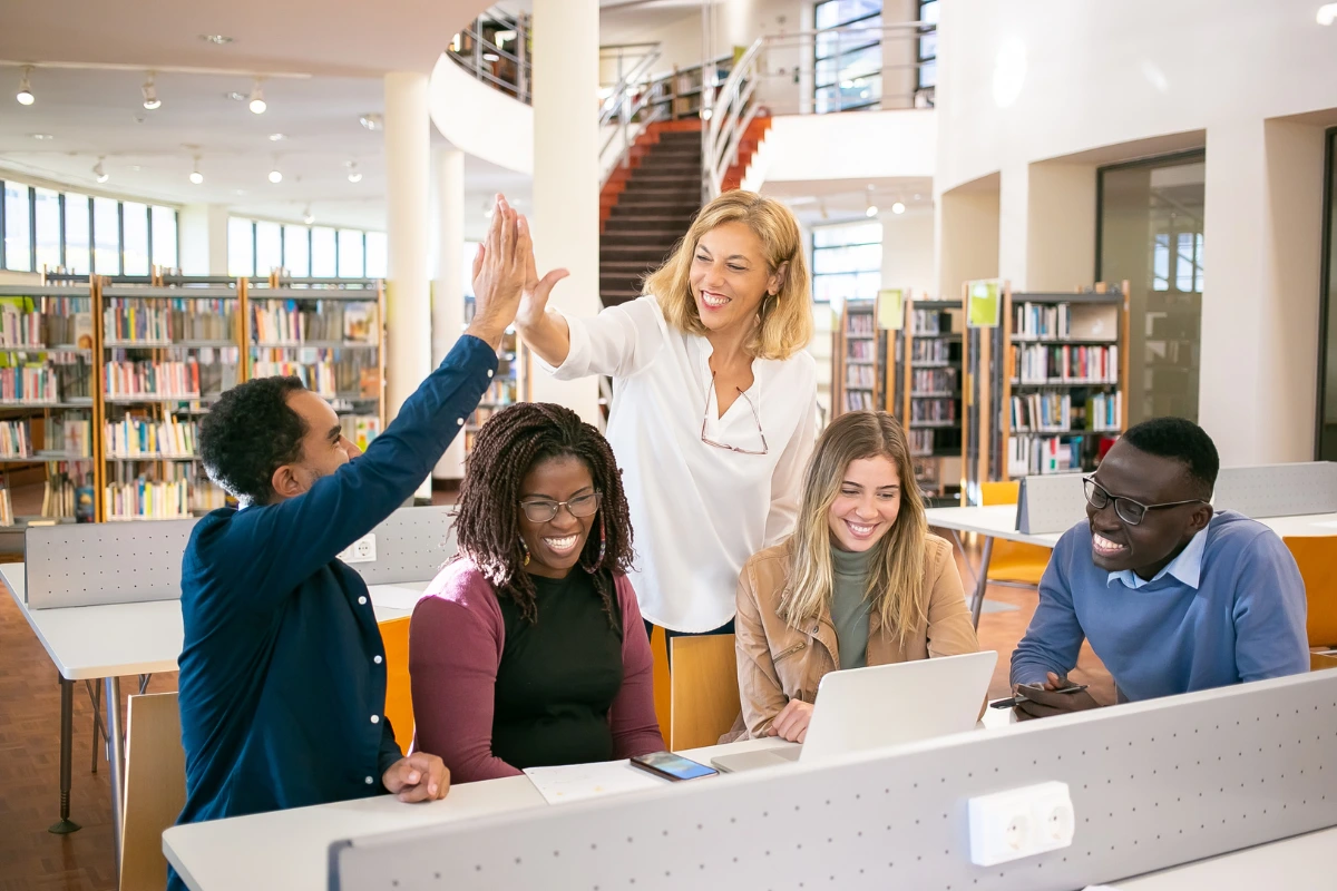 Teachers collaborating in a library, celebrating success while discussing side hustles for teachers on a laptop.