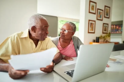 Smiling retired couple sitting at a table with a laptop and papers, discussing how to make money in retirement from home