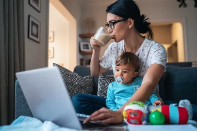 Mom holding a baby while working on a computer, exploring side hustles that actually work for busy moms to balance work and family life.