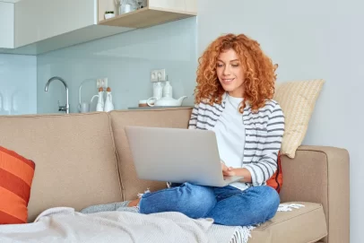 Woman sitting comfortably on a beige couch using a laptop, researching how to create a side hustle from home.