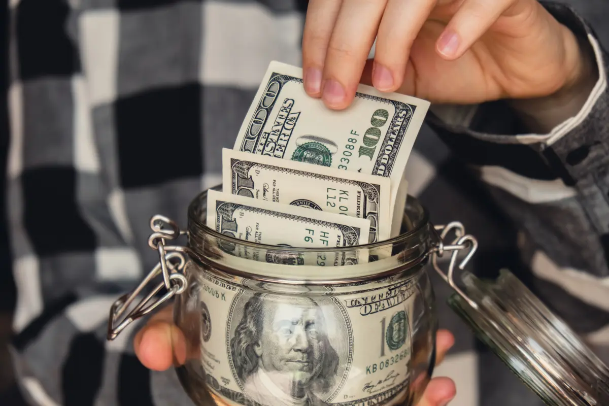Close-up of a person putting hundred-dollar bills into a glass jar, symbolizing smart saving habits and financial discipline — perfect for illustrating How to Save $10,000 in a Year Without Giving Up Everything You Love.