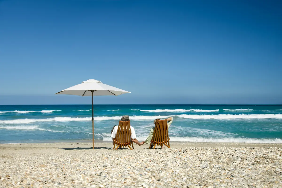 Couple relaxing on beach chairs under an umbrella, holding hands while enjoying the ocean view — symbolizing financial freedom and early retirement. Perfect for illustrating This Simple Strategy Lets You Retire 15 Years Early.