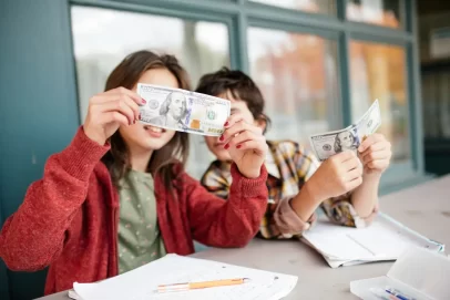 Two children sitting at a table holding dollar bills while learning a simple money habit and basic money skills.