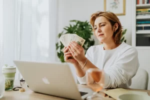 Woman counting cash while working on a laptop at home, representing unexpected ideas for a side hustle that generate extra income.