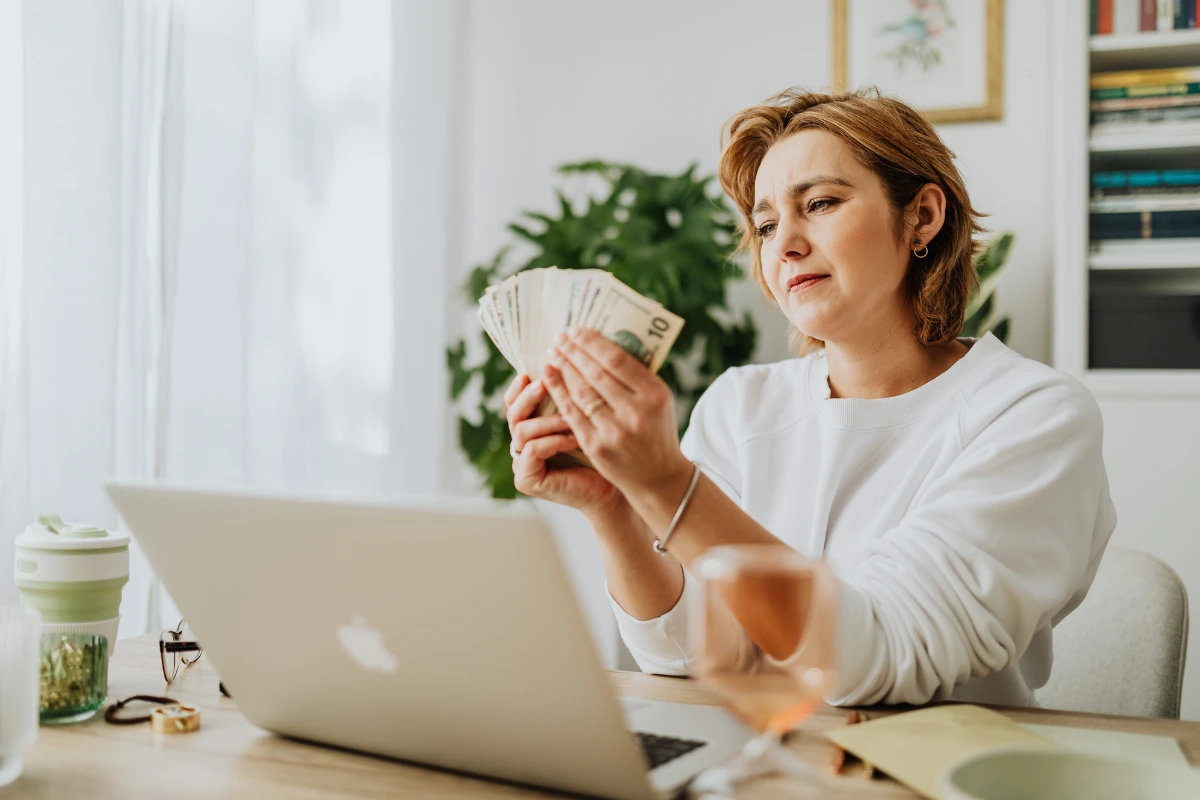 Woman counting cash while working on a laptop at home, representing unexpected ideas for a side hustle that generate extra income.